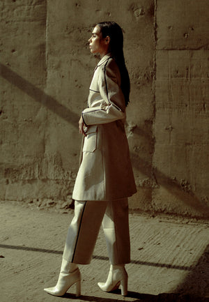 This back-pleated leather trench jacket in beige is crafted from supple faux leather and lined with luxe poly-satin. This knee-length trench coat is detailed with pleating on the back, black leather piping, cargo pockets and a leather belt for fastening. 