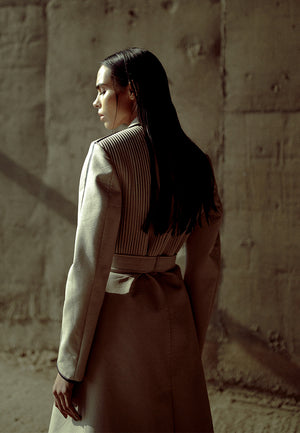 This back-pleated leather trench jacket in beige is crafted from supple faux leather and lined with luxe poly-satin. This knee-length trench coat is detailed with pleating on the back, black leather piping, cargo pockets and a leather belt for fastening. 