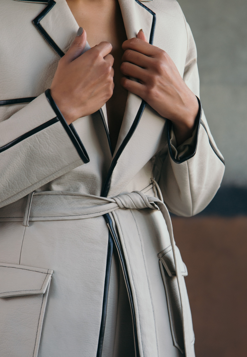 This back-pleated leather trench jacket in beige is crafted from supple rexine and lined with luxe poly-satin. This knee-length trench coat is detailed with pleating on the back, black leather piping, cargo pockets and a leather belt for fastening. Style it with a pair of pants or jeans along with boots/booties in fall and winter.