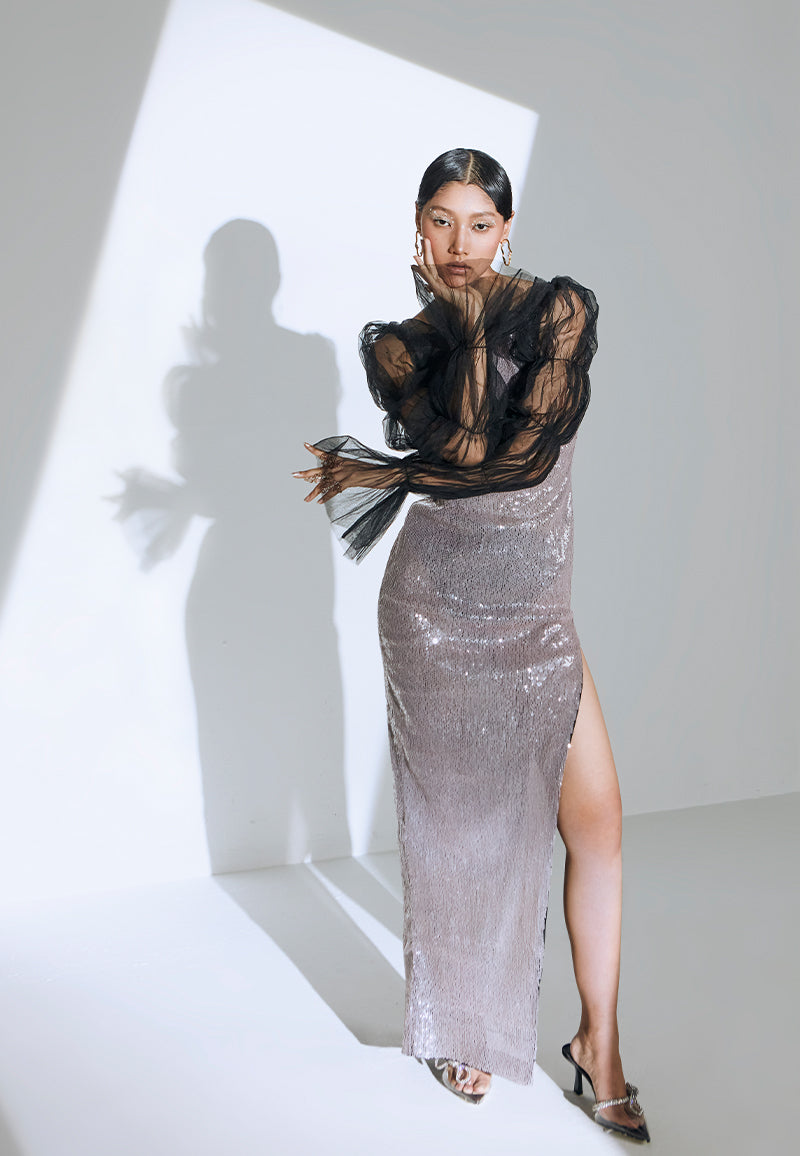 Hand-embroidered with transparent flush pink sequins, this bodycon, semi-sheer maxi dress features a boat neckline with long exaggerated, puffed sleeves in black net. The dress falls to the floor with a thigh-high slit on the side and a zip closure on the side seam. The ensemble comes with a black knit bralette and underpants. This is the perfect dress for your next big soiree. Finish the look with heeled sandals and minimal jewelry.  