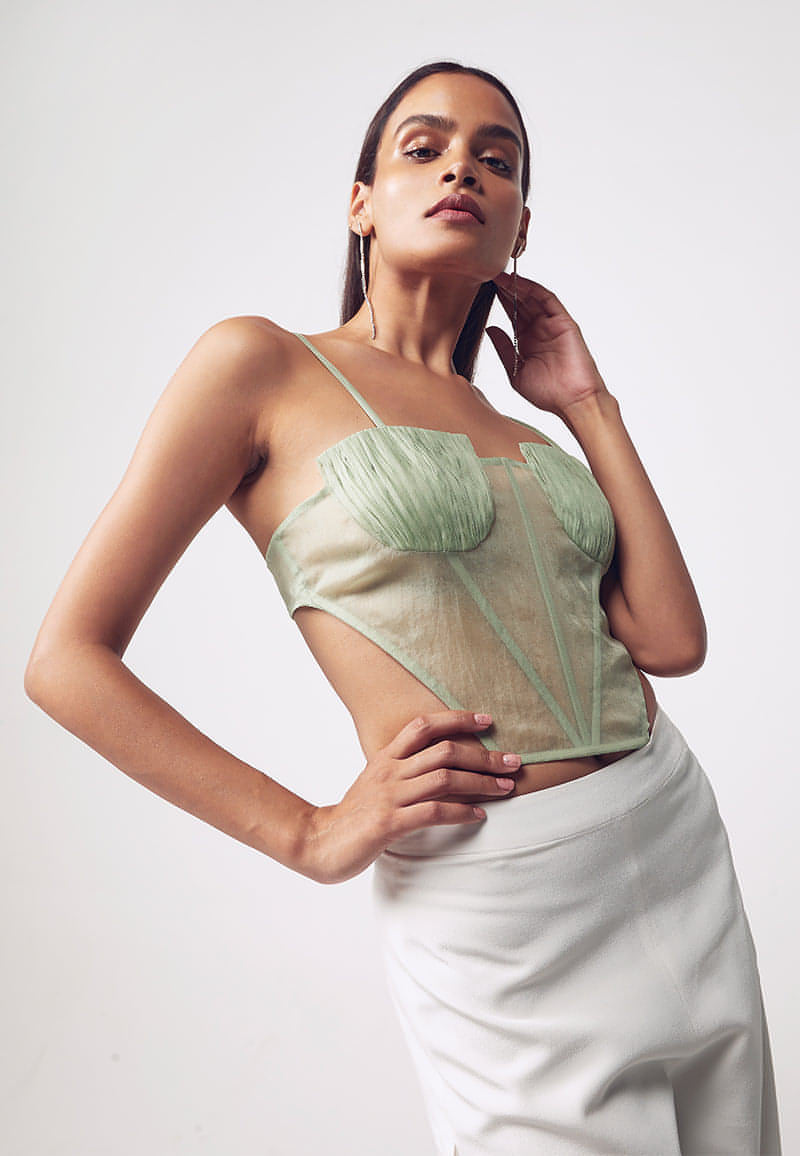 Mannat Gupta's collection of corsets, bustiers and tops. This ruched corset is made with organza in pastel green. This sleeveless, cut-out corset features ruching on the busts with a bare back. Hook and eye closures are attached at center back for seamless comfort. This minimal and versatile corsted top is perfect for any party and to deal with the summer's heat. Pair yours with one of our high-wasited pants or skirts along with heeled sandals to complete your look.  